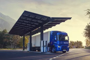 Un camion électrique bleu est en train de recharger sous une station équipée de panneaux solaires, située sur une route entourée d'arbres et de montagnes. Le ciel est dégagé avec une lumière douce du lever ou coucher du soleil, créant une ambiance paisible et écoresponsable.