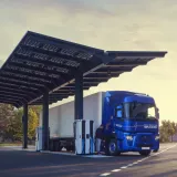 Un camion électrique bleu est en train de recharger sous une station équipée de panneaux solaires, située sur une route entourée d'arbres et de montagnes. Le ciel est dégagé avec une lumière douce du lever ou coucher du soleil, créant une ambiance paisible et écoresponsable.