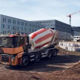 A Renault Trucks C lorry fitted with a concrete mixer is operating on a large urban construction site surrounded by cranes and modern buildings.