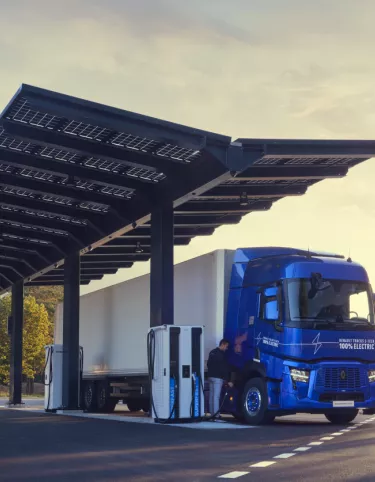 Un camion électrique bleu est en train de recharger sous une station équipée de panneaux solaires, située sur une route entourée d'arbres et de montagnes. Le ciel est dégagé avec une lumière douce du lever ou coucher du soleil, créant une ambiance paisible et écoresponsable.